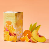 A box of Golden Coast Mixed California Gummy Bears, made with real California fruit and gluten free, sits beside orange, peach, and mango slices on a peach-colored background. The packaging displays images of fruits and gummy bears.