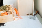The Honey Wood Box Brand features a wooden box with orange ribbon, dried flowers, and shredded paper on a white surface, alongside branding tools like a wooden panel with "honey," a metal stamp, and a utility knife.