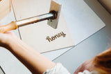 Using the Honey Wood Box Brand, a person burns "honey" in cursive onto a light wooden board, showcasing the artistry behind each brand. The workspace is clean and well-lit.