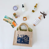 The Local Flavors Santa Barbara Traveler Tote features a jute bag filled with chocolate truffles, postcards, honey, breadsticks, olive oil, skincare items, and soap, all beautifully presented against a white background.