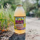 A bottle of Lori's Lavender Lemonade stands on a sunlit path amid blurred greenery. Its label showcases lavender flowers and lemons, highlighting this organic, lavender-infused, refreshing beverage.