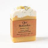 The Matilija Poppy Ojai Botanika Soap is a handcrafted, 100% vegan bar featuring a white layer with poppy seeds and dried petals, enriched with lemongrass essential oil and wrapped in a brown paper label.