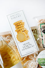 A close-up of a gift basket featuring Meyer Lemon Shortbread Cookies, a bottle of white wine, and California almond crackers nestled in shredded packing paper.