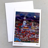 The "Moonrise over Santa Barbara Note Card" features a vibrant cityscape with a white tower, mountains, and moon, illustrated by a local artist. The colorful card comes with a crisp white envelope.