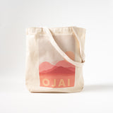 The Ojai Pink Moment Canvas Tote is a beige organic cotton bag with long handles, made in the USA. It features pink mountains and a sun graphic above bold “OJAI” lettering on the front.
