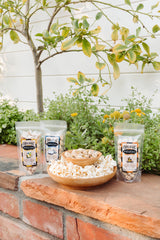 Three bags of Onion Garlic Santa Barbara Organic Pistachios (3 oz) are displayed on a brick ledge outdoors, with a bowl of pistachios and a plate of popcorn in front, set against lush greenery and a lemon tree in the background.