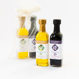 Four bottles from Santa Barbara Company, two in a sheer gift bag and two in front: one extra virgin olive oil and one fig balsamic vinegar, both with white labels and floral designs, set against a white background.