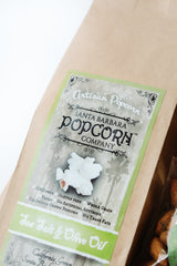 A close-up of a brown paper bag labeled Petite Sea Salt & Olive Oil Santa Barbara Popcorn, featuring California-grown, artisan popcorn that's GMO-free, gluten free, vegan, whole grain, and made without artificial ingredients.