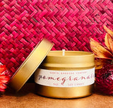The Pomegranate Travel Candle, a gold tin soy wax candle with a pomegranate scent, rests with its lid nearby on a woven pink background, surrounded by red and orange flowers.