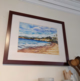 The print "Haskell's Beach and the Bacara" by Karin Shelton, featuring a beach with cliffs, ocean waves, and a partly cloudy sky, is framed and hangs above wooden kitchen utensils in a speckled ceramic container on a cream wall.