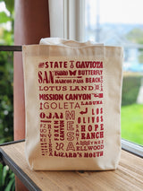 The SB Local Canvas Tote is a natural cotton bag featuring red text with local destinations like Gaviota, Lotus Land, and Hope Ranch, shown on a wooden surface by a window.
