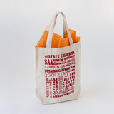 The SB Local Canvas Tote, a beige canvas bag with red local destinations text and landmarks, contains orange tissue paper and is displayed against a plain white background.