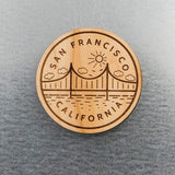 San Francisco Golden Gate Bridge Wood Magnet featuring an engraved design of the bridge, sun, clouds, and water—an ideal San Francisco gift displayed on a gray textured surface.