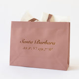 The Santa Barbara Coordinates Blush Pink Gift Tote has brown handles, white tissue paper, and "Santa Barbara 34.4° N / 119.7° W" in gold script on the front. This eco-friendly bag is shown on a plain white background.