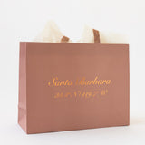 The Santa Barbara Coordinates Blush Pink Gift Tote comes with beige tissue paper and features gold script with "Santa Barbara" and 34.4°N / 119.7°W—an eco-friendly paper bag, ideal as a wedding welcome gift bag.