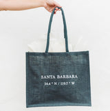 A hand holds the eco-friendly Santa Barbara Coordinates Jute Tote - Blue, featuring white text SANTA BARBARA 34.4° N / 119.7° W, against a plain white background. White tissue paper peeks out from the bag.