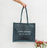 A hand holds a Miramar Santa Barbara Spa Tote labeled “Santa Barbara 34.4°N / 119.7°W” against a white wall.