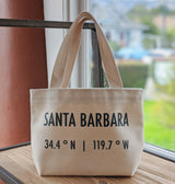The Santa Barbara Coordinates Petite Canvas Tote, a beige bag made in the USA, displays “34.4° N | 119.7° W” in black and sits on a wooden surface by a window with blurred scenery beyond.