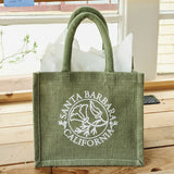 The Santa Barbara Poppy Small Tote is a green sustainable jute bag with white text and poppy floral design, reading "Santa Barbara California." The reusable gift tote sits on a wooden surface with white tissue paper peeking out.