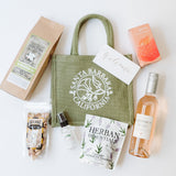 The Santa Barbara Poppy Small Tote is surrounded by snacks, a welcome card, a bottle of rosé wine, gourmet popcorn, dried oranges, pistachios, lavender towelettes, and a small spray bottle on a white background.