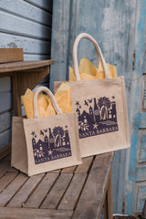 Two Santa Barbara Skyline Totes, crafted from jute cotton and featuring a cityscape design, are filled with yellow tissue paper and placed on a wooden table next to a rustic blue door and wood siding.