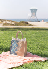 A Santa Barbara Snacks & Treats Tote with a blue geometric pattern sits on a pink blanket near the beach, ideal for carrying local snacks. In the background, a blurred lifeguard tower and ocean enhance the bright coastal vibe.