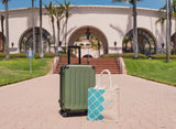 A green rolling suitcase and a Santa Barbara Snacks & Treats Tote with blue patterns are on a walkway in front of a large white building with arches—ideal for carrying local snacks on a sunny day.