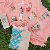 The Santa Barbara Snacks & Treats Tote rests on the grass with a pink blanket, filled with organic popcorn, dried fruit, nuts, chocolate, tea, and candy.