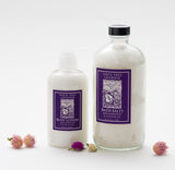 Santa Ynez Lavender Body Lotion, a hydrating organic lotion in a pump bottle with a purple label, is displayed among small pink and purple dried flowers on a white background.