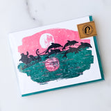 The Santa's Dolphins Letterpress Holiday Card Boxed Set features a festive design of Santa's sleigh pulled by dolphins over ocean waves, set against a large pink and white sun or moon, with a gold sticker accent in the top right corner.
