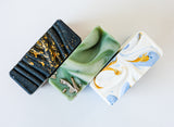 Three rectangular handmade Sea Salt Ojai Botanika Soap bars—black with gold streaks, green with metallic flakes, and white with blue and gold swirls—are arranged side by side on a white background.