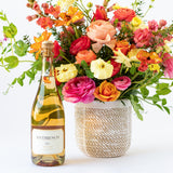 A bottle of Sparkling Brut Rosé + Flowers stands beside a woven basket filled with a vibrant bouquet, all set against a white background.