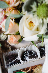 A close-up of pastel roses and greenery above a Trio of Caramels—dark, marbled chocolates and caramels in white paper wrappers—an elegant treat inspired by Santa Barbara charm.
