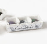 A clear box labeled "Trio of Caramels" holds three chocolate-covered caramels in white paper cups, featuring floral packaging by Santa Barbara Company.