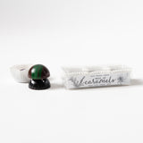 A chocolate shaped like a mushroom sits next to a clear box labeled "Trio of Caramels," containing three pieces, on a white background.