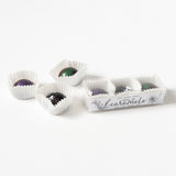 Three glossy, round chocolate covered caramels in white paper cups—two outside and one inside a clear Santa Barbara "Trio of Caramels" box—are displayed on a white background.