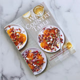 The cookbook "Wine Country Table" rests on a marble surface, showcasing vibrant salads on white plates garnished with pomegranate seeds and cheese, next to glasses of white wine.