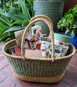 The Z - Turquoise Farmers Market & Beach Tote, filled with assorted snacks like pistachios, chocolate, and tea, sits on a brick patio surrounded by lush green plants and decorative textured pots.