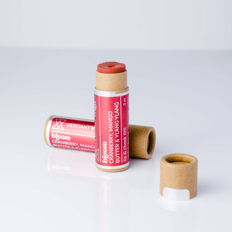 One cheek and lip balm standing with its cap off showing color of balm, and another one is placed on it side behind the standing one. Color of balm is a red orange color.