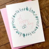 Oh Happy Day Delicate Wedding Card with floral wreath and rings on a wooden table in Santa Barbara.